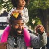A joyful African American father with curly-haired daughter on shoulders, outdoors.